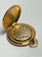 Open Bulova Accutron gold-tone hunter pocket watch showing dial, day-date window, hammered inner lid