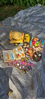 All items laid on tarp: large Tonka dump truck, small dump truck, open toy tool case with tools, plush bear, small action figures, bead necklaces, black round pouch