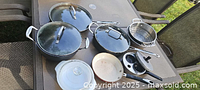 All seven cookware pieces arranged on outdoor table showing pots, pans, lids, steamer insert, egg poacher