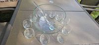 Clear glass punch bowl with glass ladle and twelve punch cups arranged around it on patio table