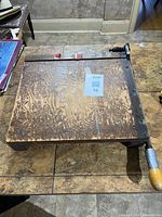 Top view of wood base guillotine paper cutter showing surface wear, red stops, blue tag, metal arm and handle