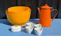 Group shot showing all items in lot: three orange bowls, orange enamel coffee pot, four white triangular holders, white textured shaker