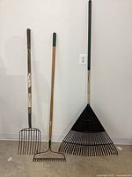 All three tools upright against wall: pitchfork, bow rake, leaf rake