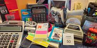 Group shot of entire lot showing calculators, tape gun, stamps, labels, cards, mesh cups, memory card