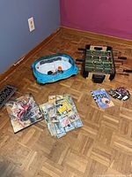 Group shot of entire lot on floor: foosball table, Beyblade arena, binder, loose cards, book, small cards