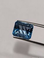 Front view of rectangular emerald-cut blue topaz held by tweezers
