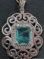 Front view of pendant showing teal gemstone, clear stone halo, scroll frame