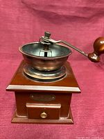 Front view of wooden coffee grinder showing drawer, brass plaque and crank handle