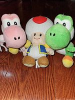 Three plush toys—pink Yoshi with scarf, Toad, green Yoshi—front view on wood floor