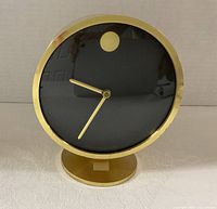 Front view of brass desk clock showing black dial, gold dot, hands, and pedestal base
