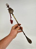 Full view of brass shoehorn held in hand showing overall length, lady finial, tassel, and spoon end