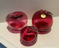Three cranberry art glass apples, front view showing sticker on one apple
