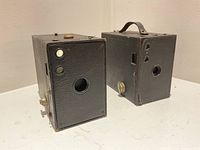 Two Kodak Brownie box cameras front view showing lenses, viewfinder windows, winding knobs and shutter levers