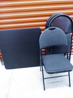 Square black folding table and stacked folding chairs