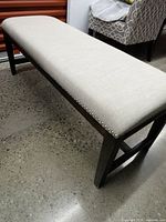 Three-quarter view of bench showing cream upholstered seat, nailhead trim, dark wood legs and stretcher on terrazzo floor
