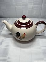 Left side view of ivory teapot showing rooster graphic, burgundy trim, lid in place