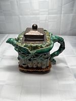 Front angle of glazed ceramic teapot showing leaf/bamboo motif and pewter lid
