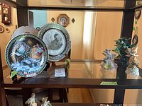 Overall view of three plates, porcelain figurines and faux plant on display shelf
