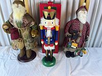 Three Christmas figures: tapestry-coat Santa, wooden nutcracker, burgundy cloth Santa; red nutcracker box visible