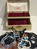 Cream Texol jewelry box open with red velvet lining and pile of beaded jewellery in front
