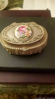 Angled view of the ornate gold-tone oval trinket box showing raised decoration and porcelain floral insert