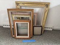 Stack of eight empty picture frames in assorted sizes and finishes leaning against wall on carpet