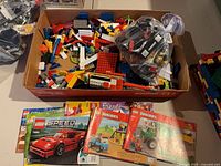 Open box showing assorted LEGO pieces, sealed bag of black elements, instruction manuals visible