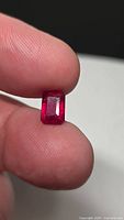Rectangular emerald-cut ruby held between fingers, face-up view showing color and cut
