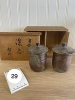 Two Raku cups with lids fronted by storage box and lot card