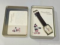 Open tin showing wristwatch, paper insert, brown strap, gold-tone case