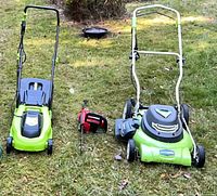 Two green electric lawn mowers and one red chainsaw on grass