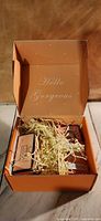 Open peach colored gift box with lid text 'Hello Gorgeous' showing packed bath items