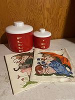 Two red canisters and two cross-stitch panels visible