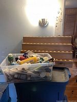 Bin of assorted craft paints, brushes on storage tote with wooden rack behind
