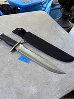 Knife and sheath displayed on table showing full length, serrated spine, handle detail, sheath beside