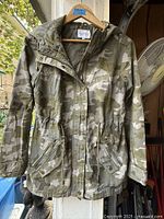 Front view of camouflage jacket on hanger showing hood, zipper, pockets and overall condition