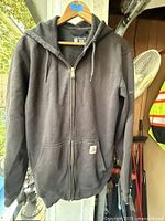 Front view of black Carhartt hooded zip sweatshirt on hanger showing pockets, zipper, hood, and logo patch