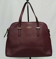 Front view of burgundy Kate Spade Cedar Street Maise satchel, showing logo, top handles, and gold hardware