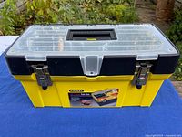 Front view of yellow Stanley tool box, lid closed, showing metal latches and clear top compartment
