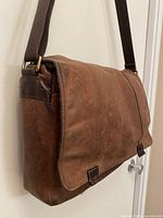 Left side view of brown leather messenger bag showing flap and strap attachment