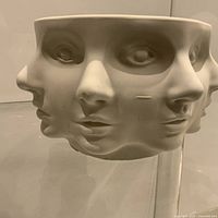 Front view of unglazed pedestal bowl with multiple abstract faces; shows overall form