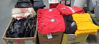 overview of three boxes containing black duffle bags, cotton tote bags, and stacks of red and yellow T-shirts