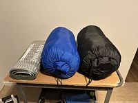 Three rolled items: two sleeping bags in stuff sacks (blue, black) and one silver Therm-a-Rest foam pad