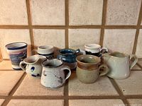 Group of eight handmade ceramic mugs and creamers on tiled surface