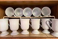 Front view showing all six white pedestal Irish coffee mugs on lower shelf