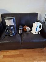 All items together on sofa: coffee maker, travel mug, teapot, glass mug with infuser, electric kettle