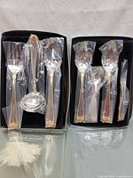 Two Heritage Silversmiths boxed sets opened to show gold-accented flatware pieces in plastic sleeves