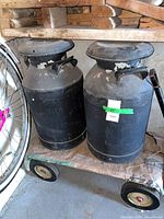 Two black metal dairy milk cans on dolly showing size and overall wear