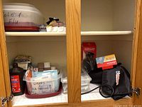 Cabinet shelf showing hydrogen peroxide bottle, clear storage bins labeled wraps/bandages, adhesive bandage box, black blood pressure cuff case, Mueller brace box, Sunbeam heating pad box