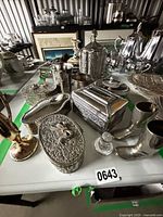 Group shot showing all silver-tone boxes, tumblers, boot vessels, bud vase, and perfume bottle within green border